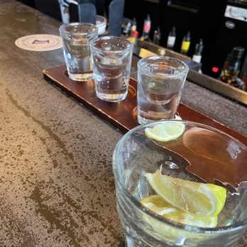 Chili Lime - Three shot glasses filled with clear liquid are arranged on a wooden tray on a bar counter. In the foreground, a glass with lemon wedges is visible. The background shows a bar area with bottles and bar equipment, indicating a bar or pub setting. - Tacos Near Me - Champlin, Minnesota