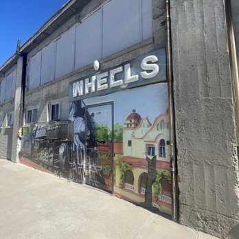 Wheels Museum - A mural on a building wall depicts a train, a church, and a cat, with the word "WHEELS" in large letters above. The mural is painted on a concrete wall near a sidewalk. - Museums Near Me - Albuquerque, New Mexico