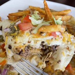 Smothered Tortilla Burger - A tortilla burger topped with shredded cheese, lettuce, tomato, and French fries on a white plate, with a glass of water in the background.