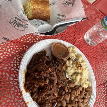 Willards Real Pit BBQ - Burnt Ends - A disposable white plate contains shredded barbecue meat with sauce, baked beans, and mashed potatoes topped with gravy. A small plastic cup of barbecue sauce is placed on the plate. A slice of cake with a golden-brown crust is wrapped in branded paper, with a plastic fork resting on it. A bottle of water is positioned nearby on a red metal table with a perforated pattern, suggesting an outdoor dining setting. - Barbeque Near Me - Reston, Virginia