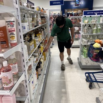 Marshalls - A person wearing a green shirt, black shorts, and sneakers is shopping in a retail store, reaching for a product on the shelf in the personal care aisle. The store has multiple shelves stocked with bottles and containers of various personal care items. The background shows signage for different sections, including 'intimates' and other product categories, with overhead lighting illuminating the area. - Department Stores Near Me - Costa Mesa, California