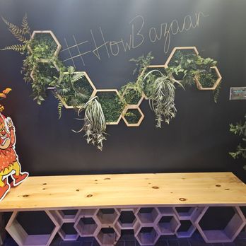 Bristol Bazaar - Crafty entrance spot. - The image shows a wall decorated with multiple hexagonal planters containing various green plants, arranged in a honeycomb pattern. Above the planters, the hashtag #HowBazar is written in chalk or marker. In front of the wall, there is a wooden bench with a geometric honeycomb design on its sides. To the left, a cartoon character cutout is visible, and to the right, a small electronic keypad is mounted on the wall. The setting appears to be an indoor space at Bristol Bazaar, supporting artisans and community activities. - Public Markets Near Me - Bristol, Connecticut