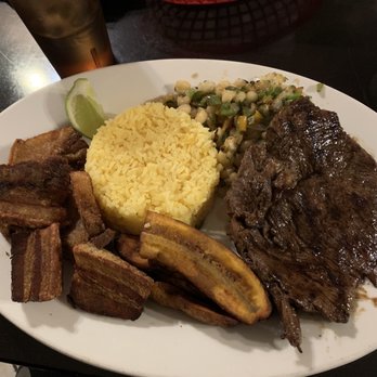 La Cascada - Roast pork, Fried pork, rice, plantains, corn/potato mix and beans on the side! - A plate featuring yellow rice, fried plantains, beans, and a corn and green onion salad, with a lime wedge, served with a beef steak. - Latin American Near Me - Peekskill, New York