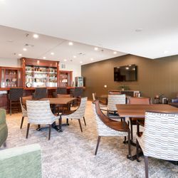 Vista Prairie at Copperleaf - A warm and inviting social lounge called "Suds." It features a dark wood bar with glass shelving, several bistro tables with ...