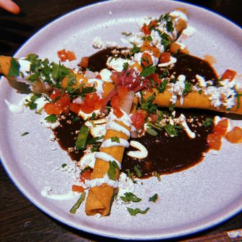 Anafre - A white plate holds two taquitos topped with dark sauce, sour cream, crumbled cheese, chopped tomatoes, and cilantro. The dish is garnished with additional cilantro and sauce drizzles, presented on a dark wooden table. - Mexican Near Me - Washington, DC, District of Columbia