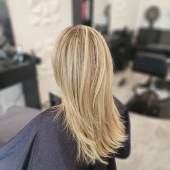 Salon Luxe - The image shows a woman with blonde, layered hair sitting in a salon chair, facing away from the camera. The salon interior includes styling stations, mirrors, and salon equipment, indicating a professional hair salon setting. The woman is wearing a dark cape, typical for hair treatments. - Hair Salons Near Me - Lindenhurst , Illinois