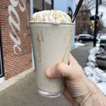 DMK Burger Bar - SOTM Apple Pie Shake - A person holds a tall glass of milkshake topped with whipped cream and caramel drizzle. The background shows a brick wall with a partially visible sign, a sidewalk, and some snow on the ground. The scene is outdoors in an urban setting with parked cars and street signs visible. - Burgers Near Me - Chicago, Illinois