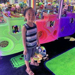 A young girl with short dark hair, wearing a striped sleeveless top and shorts, stands in front of claw machines filled with plush toys at an arcade. She holds several plush toys in her hand, and the background features brightly lit claw machines with various stuffed animals. The arcade has neon lighting, colorful machines, and a game area with a black carpet and green accents.