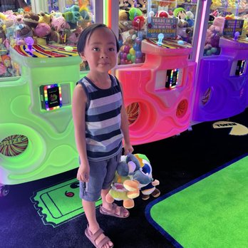 Fun Zone Arcade - A young girl with short dark hair, wearing a striped sleeveless top and shorts, stands in front of claw machines filled with plush toys at an arcade. She holds several plush toys in her hand, and the background features brightly lit claw machines with various stuffed animals. The arcade has neon lighting, colorful machines, and a game area with a black carpet and green accents. - Arcades Near Me - Vacaville, California