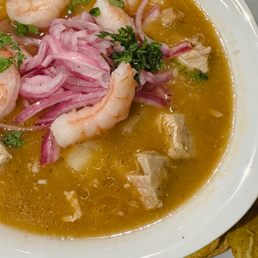 Encebollado - A bowl of Ecuadorian fish soup, encebollado de pescado, contains shrimp, chopped red onions, and fresh herbs in a yellowish broth. The dish is served in a white bowl on a dark wooden table, with some potato chips visible at the edge of the plate. The soup includes chunks of fish and garnishes of cilantro and parsley.
