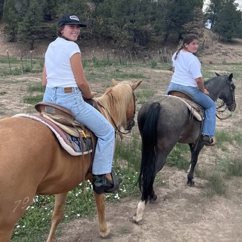 Mesa Verde Stables - Two girls are riding horses on a dirt trail surrounded by trees and open land, wearing casual clothing and helmets. - Horseback Riding Near Me - Mancos, Colorado
