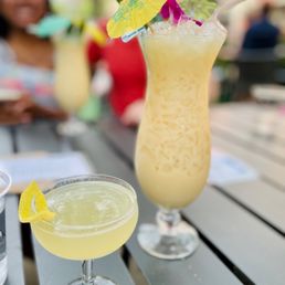 Virgin Pina Colada - Two cocktails with yellowish drinks are on a table outdoors, one in a tall hurricane glass with a paper umbrella, and the other in a coupe glass with a lemon garnish.