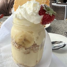 Banana cream pudding - A layered dessert served in a glass jar, topped with whipped cream, fresh strawberries, and a large cookie, placed on a table.