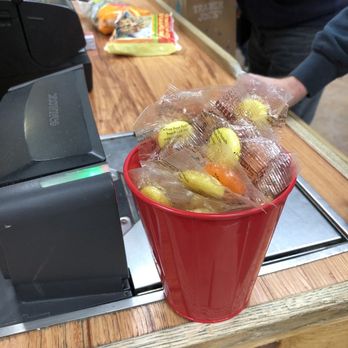 Trader Joe's - Free lollipops if you find Mochi and Pickles - A red plastic cup on a store checkout counter contains individually wrapped candies, including yellow and orange pieces. The counter has a wooden surface, and a cash register is visible on the left. In the background, there are snack items and a person wearing a dark jacket and gloves, indicating a retail environment. - Grocery Near Me - Danbury, Connecticut