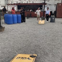 Corn Hole. - An outdoor bar area features cornhole game boards on gravel ground, with several people standing near a bar counter and blue barrels.