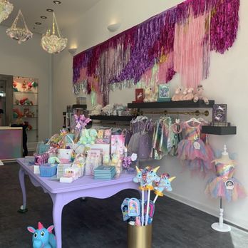 The Unicorn Store - Store merchandise - The store features a variety of children's clothing, plush toys, and accessories displayed on shelves and a lavender-colored table. A large, colorful, fringed wall hanging in shades of pink, purple, and gold decorates the wall behind the display. Mannequins dressed in rainbow-colored tutus are positioned on the right side. The store has a high ceiling with hanging chandeliers and wall-mounted lights, creating a bright and playful atmosphere. - Children's Clothing Near Me - Pasadena, California