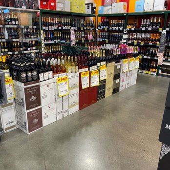 BevMo! - The image shows a section of a liquor store with shelves filled with wine bottles, including sparkling and still wines. There are boxes of wine stacked on tables and shelves, with sale signs indicating discounts. The store has a concrete floor with visible cracks and colorful wall stripes in red and yellow. The display is organized with bottles grouped by type and brand, and promotional signs are attached to the tables. - Beer, Wine & Spirits Near Me - Temecula, California
