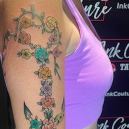 The image shows a person's upper arm with a tattoo of a floral design. The tattoo includes multiple flowers in shades of yellow, pink, blue, and purple, connected by green leaves and vines. The person is wearing a purple sleeveless top, and the background features a tattoo studio setting with signage. The tattoo appears detailed and vibrant.