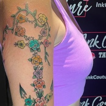 Ink Couture - The image shows a person's upper arm with a tattoo of a floral design. The tattoo includes multiple flowers in shades of yellow, pink, blue, and purple, connected by green leaves and vines. The person is wearing a purple sleeveless top, and the background features a tattoo studio setting with signage. The tattoo appears detailed and vibrant. - Tattoo Near Me - Austin, Texas