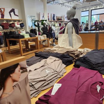 Athleta - Clothing items such as t-shirts and sweatshirts are folded on tables in a retail store, with mannequins dressed in activewear and casual clothing in the background. - Sports Wear Near Me - Avon, Connecticut