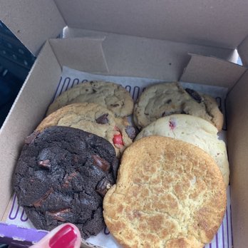 Insomnia Cookies - A person with red nail polish holds a cardboard box filled with six cookies, including chocolate chip, double chocolate, and sugar cookies. The cookies are arranged in two rows inside the box, which has a purple and white paper liner. The background shows part of a vehicle interior, indicating the cookies are being held in a car. - Desserts Near Me - Albuquerque, New Mexico