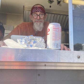 Simpson's BBQ - Simpson's BBQ server, offering a brisket taco and drink. - A man with glasses, a bandana, and a beard is smiling behind a food truck window, with food and a drink on the counter. - Food Trucks Near Me - Manchaca, Texas