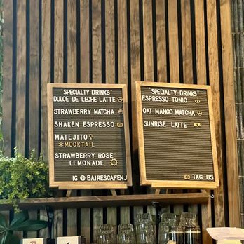 Cafe Baires - Menu as of November 2025 - Two black felt letter boards with wooden frames are mounted on a wooden wall at Cafe Baires. The left board lists specialty drinks such as dulce de leche latte, strawberry matcha, shaken espresso, matejito, strawberry rose lemonade, and an Instagram handle. The right board features espresso tonic, oat mango matcha, and sunrise latte, with icons indicating flavors. Below the boards, there are glass cups, coffee bags, and takeaway cups on a shelf. - Cafes Near Me - Hackensack, New Jersey