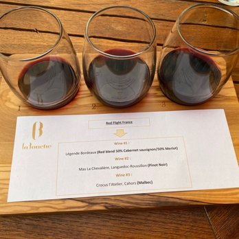 La Bouche - Rouge French wine flight - Three glasses of red wine are arranged on a wooden tasting board, each filled with different amounts of wine, accompanied by a wine flight menu from La Bouche. - Wine Bars Near Me - Denver, Colorado