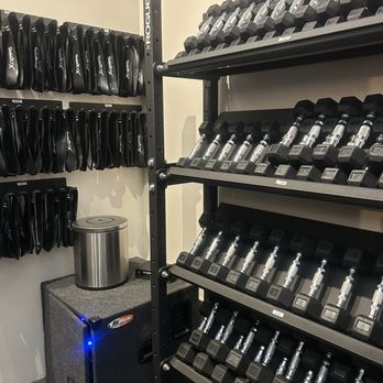 The Foundry - A fitness storage room contains multiple shelves of dumbbells organized by weight, with labels visible. On the left wall, there are several racks holding weightlifting belts. A small refrigerator is placed on a carpeted stand in the corner, with a metallic trash can on top. The room has plain walls and a neutral floor, designed for storing workout equipment. - Yoga Near Me - Mesa, Arizona