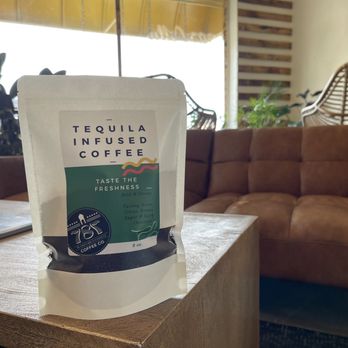 787 Coffee - A view of the coffee atmosphere and tequila infused coffee beans. - A package of tequila-infused coffee labeled 'Taste the Freshness' is positioned on a wooden table inside a cafe. The package is white with a green and black label, and it indicates an 8 oz size. The background includes a brown sofa, potted plants, and large windows allowing natural light, creating a relaxed cafe atmosphere. - Coffee & Tea Near Me - El Paso, Texas