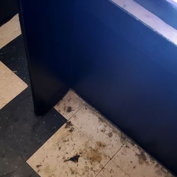 Pleasure World - This place needs a MAJOR CLEAN UP LIKE SO BAD!!! Need chairs too in the booths. - The image shows a section of tiled flooring with black and white tiles, some of which are stained and dirty, with visible debris and grime. A blue booth or counter is partially visible in the background, with a lit display or screen nearby. The area appears to be in need of cleaning and maintenance. - Adult Shops Near Me - Phoenix, Arizona