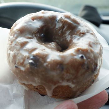 Kim’s Donuts - Blueberry - A close-up of a glazed blueberry donut with icing and blueberries on top, held in a person's hand inside a vehicle. The background shows part of the car's dashboard and windshield, indicating the photo was taken while seated in a car. The donut appears freshly made and is presented on a white paper napkin. - Donuts Near Me - Lowell, Massachusetts