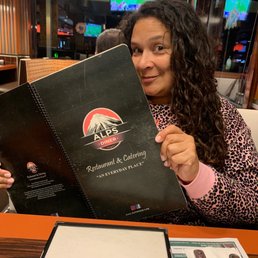 Me in love with the idea of having cream of turkey! - A woman with curly dark hair, wearing a pink leopard-print top, is seated at a table in a restaurant, holding a black Alps Diner menu prominently in front of her. The restaurant interior features wood paneling, and multiple TV screens displaying sports are visible in the background. The table has a paper placemat and a menu holder, indicating a casual dining environment.