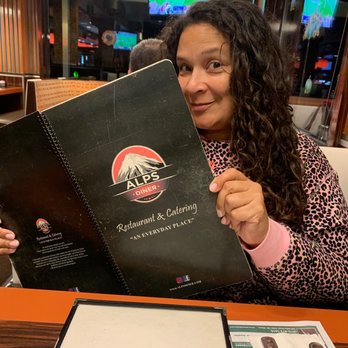 Alps Diner - Me in love with the idea of having cream of turkey! - A woman with curly dark hair, wearing a pink leopard-print top, is seated at a table in a restaurant, holding a black Alps Diner menu prominently in front of her. The restaurant interior features wood paneling, and multiple TV screens displaying sports are visible in the background. The table has a paper placemat and a menu holder, indicating a casual dining environment. - Diners Near Me - Wayne, New Jersey