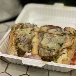 Beef Brisket Sandwiches - A beef brisket sandwich features sliced beef, melted cheese, onions, and herbs inside a sandwich bun. The sandwich is served in a white, rectangular takeout container on a tiled surface. The image also shows a red canned beverage partially visible on the left side. The sandwich appears to be freshly prepared with toppings and cheese visibly melted over the beef.