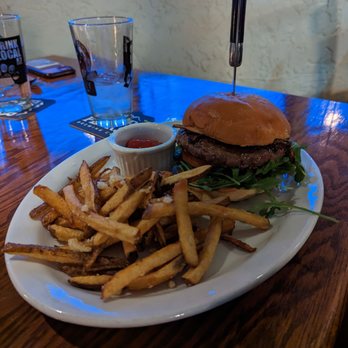 Il Vinaio - Antelope burger with fries - A plate with a burger featuring a beef patty, lettuce, and a bun is placed on a wooden table. The burger has a knife inserted through the top bun. Accompanying the burger are French fries and a small container of ketchup. In the background, there are two empty glasses and a coaster, indicating a dining setting in a restaurant. - New American Near Me - Mesa, Arizona