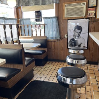 Jefferson Boulevard Diner - Seating area - The image shows the interior of a classic American diner with booth seating that has black cushions and wooden frames. Checkered curtains adorn the windows, allowing natural light to enter. The walls are covered with wood paneling and decorated with a large Elvis Presley poster, framed photographs, and a green chalkboard with a golf club illustration. An air conditioning unit is mounted on the wall, and there are three black and chrome bar stools in the foreground. The floor is tiled with small, square, brown tiles, and a black mat is placed near the booth. - Breakfast & Brunch Near Me - Warwick, Rhode Island