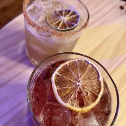 Tamarind Margarita & Blackberry Sour - Two cocktails with lemon slices and ice are shown on a light-colored wooden table. One drink has a salted rim and a light color, while the other is darker with visible bubbles and black peppercorns. The drinks are served in short glasses, and the setting appears to be in a bar or restaurant environment.
