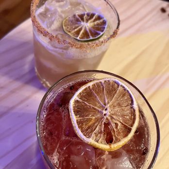 Downshift Brewing: Outpost 1706 - Tamarind Margarita & Blackberry Sour - Two cocktails with lemon slices and ice are shown on a light-colored wooden table. One drink has a salted rim and a light color, while the other is darker with visible bubbles and black peppercorns. The drinks are served in short glasses, and the setting appears to be in a bar or restaurant environment. - Beer, Wine & Spirits Near Me - Albuquerque, New Mexico