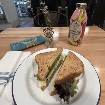 The Corner Counter - Prairie Breeze sandwich - A sandwich with lettuce, cheese, and other ingredients is presented on a white plate with a blue rim, placed on a wooden table in a cafe. The sandwich is cut in half, showing the filling inside. A bottle of raspberry lemonade is positioned behind the plate. The background includes a vase with dried flowers, a menu, and cafe seating with chairs and a counter. - Sandwiches Near Me - Red Hook, New York
