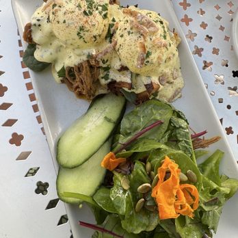 Coronet Cafe - Eggs Benedict with hollandaise sauce, shredded meat, and garnishes, served with a side salad and cucumber slices on a rectangular plate. - Cafes Near Me - Tucson, Arizona