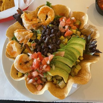Metate - Taco Salad with Grilled Shrimp - A taco salad presented in a large, crispy tortilla bowl features grilled shrimp, sliced avocado, black beans, diced tomatoes, chopped onions, and corn. The dish is arranged with fresh ingredients, highlighting Mexican cuisine elements, and is served on a white plate with additional sides visible nearby. - Mexican Near Me - Bronx, New York