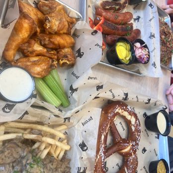 Prost Brewing - Deliciousness to share - The photo features multiple dishes on a table, including a basket of chicken wings with celery sticks and dipping sauce, a tray of assorted sausages with mustard, pickles, and onions, a large pretzel with salt, and a plate of steak with fries and gravy. Small containers of sauces and condiments are also visible, arranged on parchment paper and a wooden table surface. - Breweries Near Me - Northglenn, Colorado