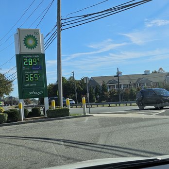 bp - October 2025 - The image features a BP gas station sign displaying fuel prices, with regular gasoline at $2.89 per gallon and diesel at $3.69 per gallon. The sign is situated on a paved area with small bushes and yellow safety posts. In the background, there are residential houses, trees, and utility poles under a partly cloudy blue sky. The scene appears to be taken from inside a vehicle, with part of the dashboard visible at the bottom of the image. - Gas Stations Near Me - West Babylon, New York