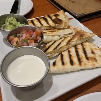 La Malinche - Quesadilla de Camarones - A rectangular white plate holds three grilled quesadilla de camarones slices with charred grill marks. Accompanying the quesadillas are small metal cups containing guacamole, pico de gallo, and sour cream. The dish is set on a wooden table with a menu and utensils visible in the background, indicating a dining setting at La Malinche in Silver Spring, MD. - Tapas Bars Near Me - Silver Spring, Maryland