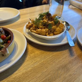 True Food Kitchen - Cauliflower - A cauliflower dish with herbs and garnishes is presented on a white plate at a restaurant table. The dish appears to be seasoned and topped with fresh herbs like dill and mint. Nearby, there is a glass of water, a person holding a drink, and other plates of food. The setting includes a wooden table and a napkin, indicating a dining environment. - New American Near Me - Oakbrook, Illinois