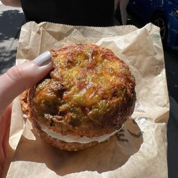 Green Chili and cheese bagel with plain cream cheese - A bagel topped with green chili, cheese, and melted cheese, held by a person with painted nails, with a person and cars in the background.