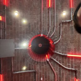 Ceiling with exposed metal pipes arranged in various directions, illuminated by red neon lights. A circular black vent or fixture is centrally located on a brick wall, with small lights and vents visible around. The setting appears to be an interior space with industrial or modern design elements.