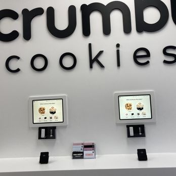 Crumbl - Delafield - a display of crumbl cookies - Two digital ordering tablets are mounted on a white wall inside Crumbl Cookies, each displaying menu options for cookie selection. The store's name, 'crumbl cookies,' is prominently displayed in large black letters above the tablets. The setup appears to be part of a self-service ordering station, with a small black device and a sign indicating job openings placed on the white counter below. - Bakeries Near Me - Delafield, Wisconsin