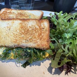 Breakfast sandwich - A breakfast sandwich featuring toasted bread with a filling of greens and melted cheese is placed on a plate. Next to the sandwich, there is a side salad composed of mixed leafy greens. The plate is set on a table with glasses and other items visible in the background.